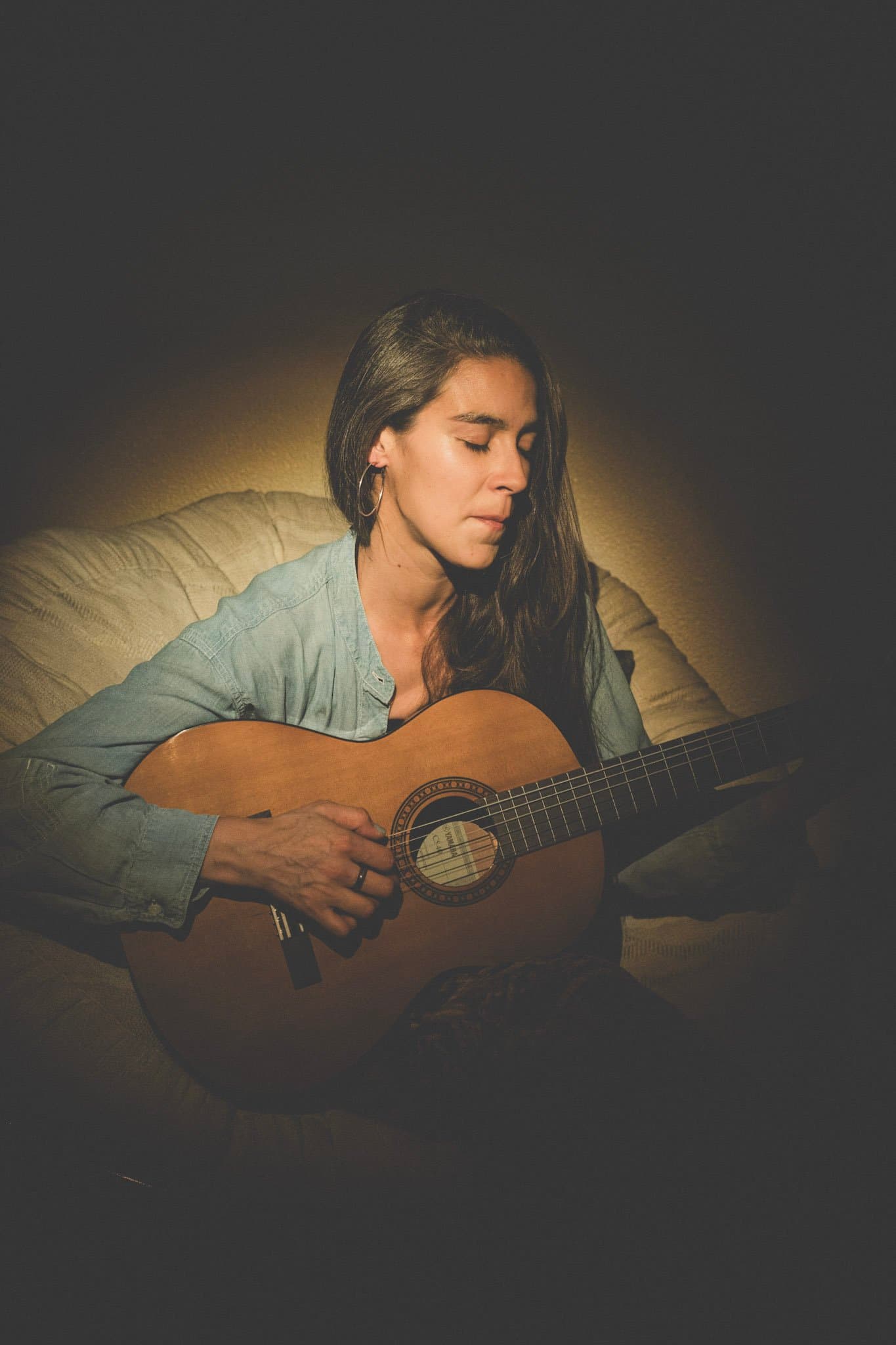 A woman with long dark hair, wearing a light blue denim shirt, sits on a beige couch playing an acoustic guitar, with her eyes closed and a serene expression.