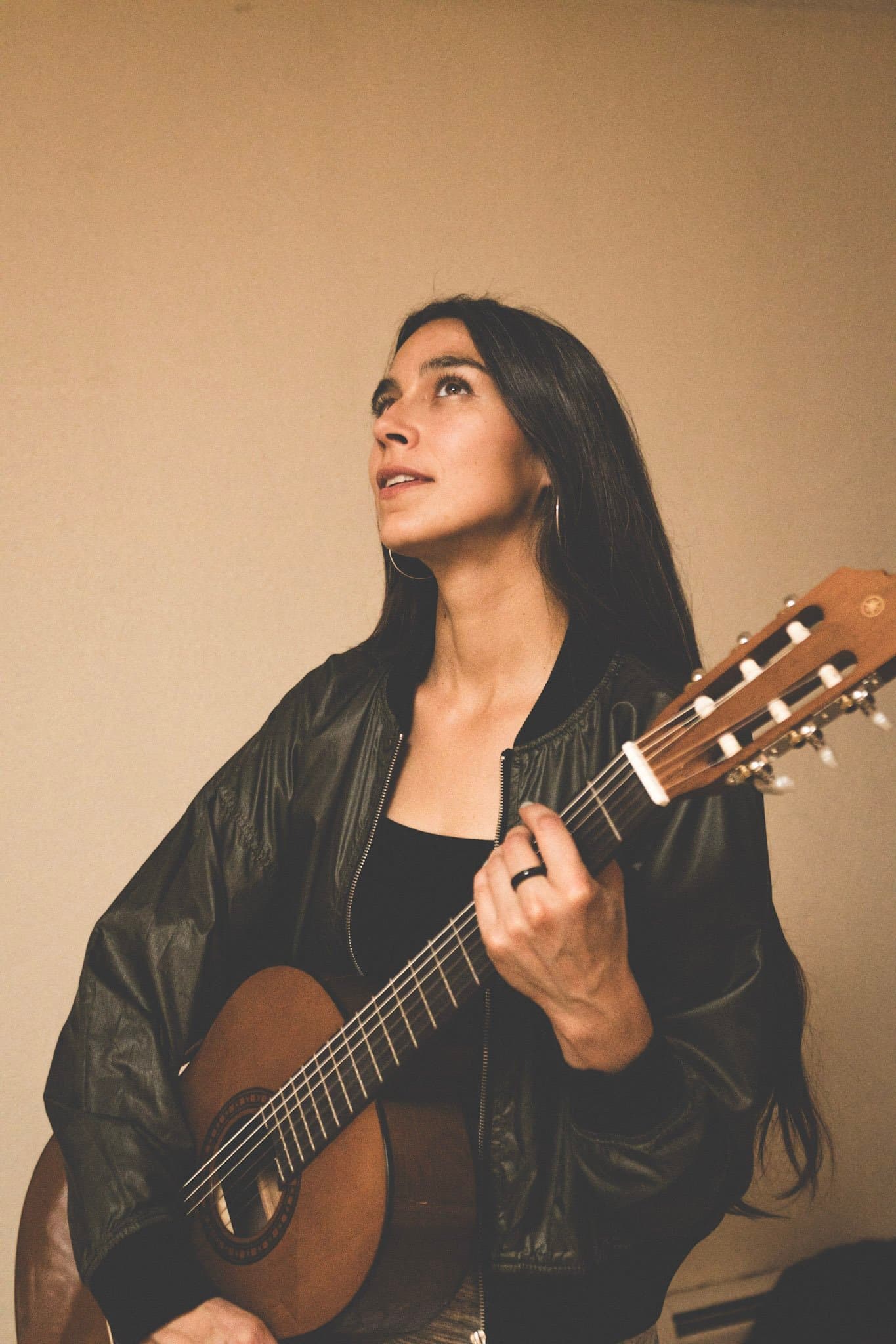 A woman with long dark hair is playing an acoustic guitar and looking upwards, wearing a black jacket and hoop earrings, standing against a plain wall.